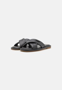 Sorte sorte sandaler i svart skinn med kryssdesign, flat såle og teksturert innside. Yttersålen er beige for kontrast.