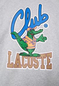 Felpa grigia con una grande grafica colorata di un coccodrillo verde che indossa una maglietta arancione e un berretto, insieme a scritte blu e marroni.