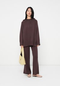 Femme portant un sweat à capuche loose violet foncé et un pantalon large assorti, tenant un sac à main beige clair et portant des ballerines rose pâle.