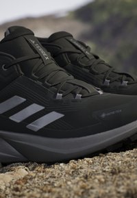 Czarne buty trekkingowe mają teksturowaną syntetyczną cholewkę, wyraźne szare akcenty i solidną podeszwę, zaprojektowane z myślą o wytrzymałości na zewnątrz.