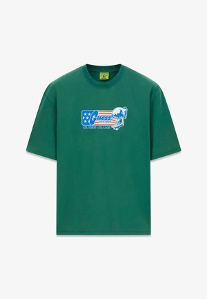 Grünes T-Shirt mit kurzen Ärmeln, blau-rotem "Guess Jeans"-Logo und einer Grafik von einer Person, die auf dem Brustbereich Skateboard fährt.