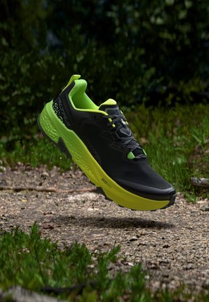 Scarpa da corsa nera e verde neon sospesa sopra un sentiero di ghiaia con erba e fogliame sullo sfondo.