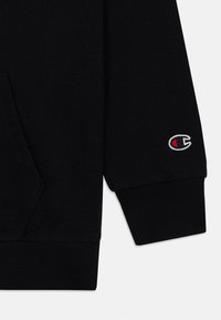 Sweatshirt noir au tissu doux, doté d'une poche kangourou et d'un petit logo brodé en blanc et rouge près du poignet.