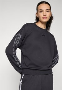Donna con felpa nera Adidas con strisce ornate sulle maniche e pantaloni neri abbinati con strisce bianche sui lati, in piedi di fronte a uno sfondo semplice.
