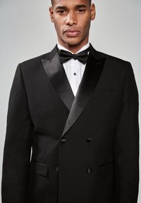 Sort smoking med satin revers, dobbeltradet design og en hvid skjorte med knappet krave. Butterfly inkluderet.