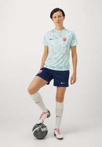 Nike damfotbollskit bestående av en ljusblå tröja med geometriskt mönster, marinblå shorts, vita strumpor och röda detaljer på skorna, tillsammans med en svart boll.