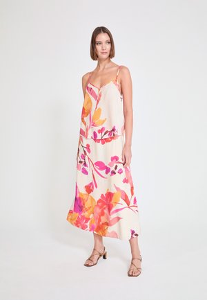 Mujer con top sin mangas y falda larga con grandes estampados florales en rosa brillante, naranja y morado, de pie frente a un fondo claro y liso.