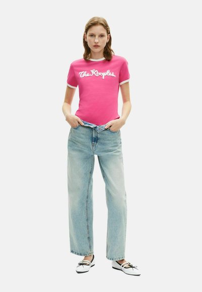 Roze T-shirt met korte mouwen en witte opdruk, gecombineerd met lichtblauwe broek met wijde pijpen. Het model draagt zwart-witte schoenen.