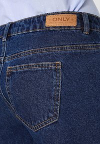 Tmavě modré džíny z denimu s oranžovým prošíváním, s koženou značkou na pásku a zadní kapsou s klasickým střihem.