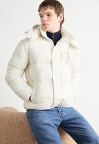 Kremfarget pufferjakke med hette, med glidelås foran og beige logo. Myk tekstur, quiltet design, kombinert med blå jeans.