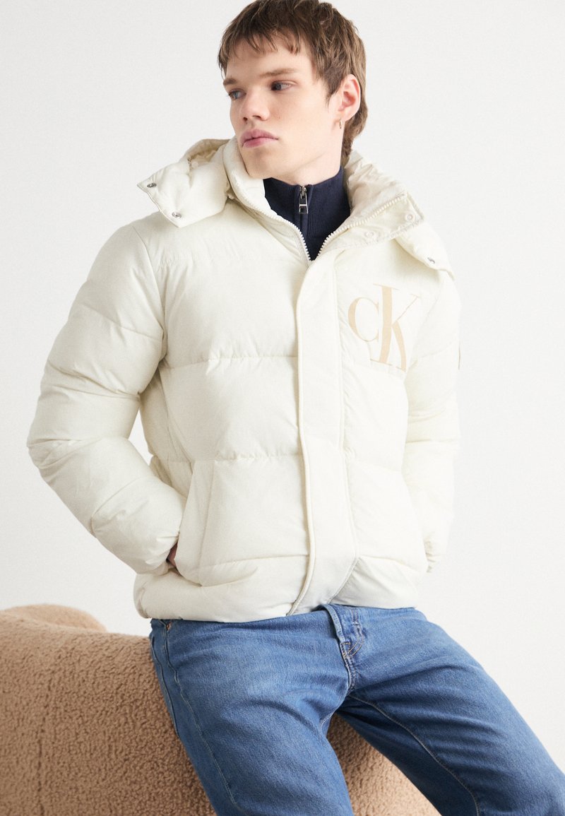 Kremfarget pufferjakke med hette, med glidelås foran og beige logo. Myk tekstur, quiltet design, kombinert med blå jeans.