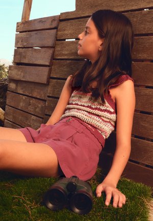 Djevojka u pletenom topu i kratkim hlačama sjedi naslonjena na drveni zid na otvorenom, sa dvogledom na travi pored sebe, gleda udesno.