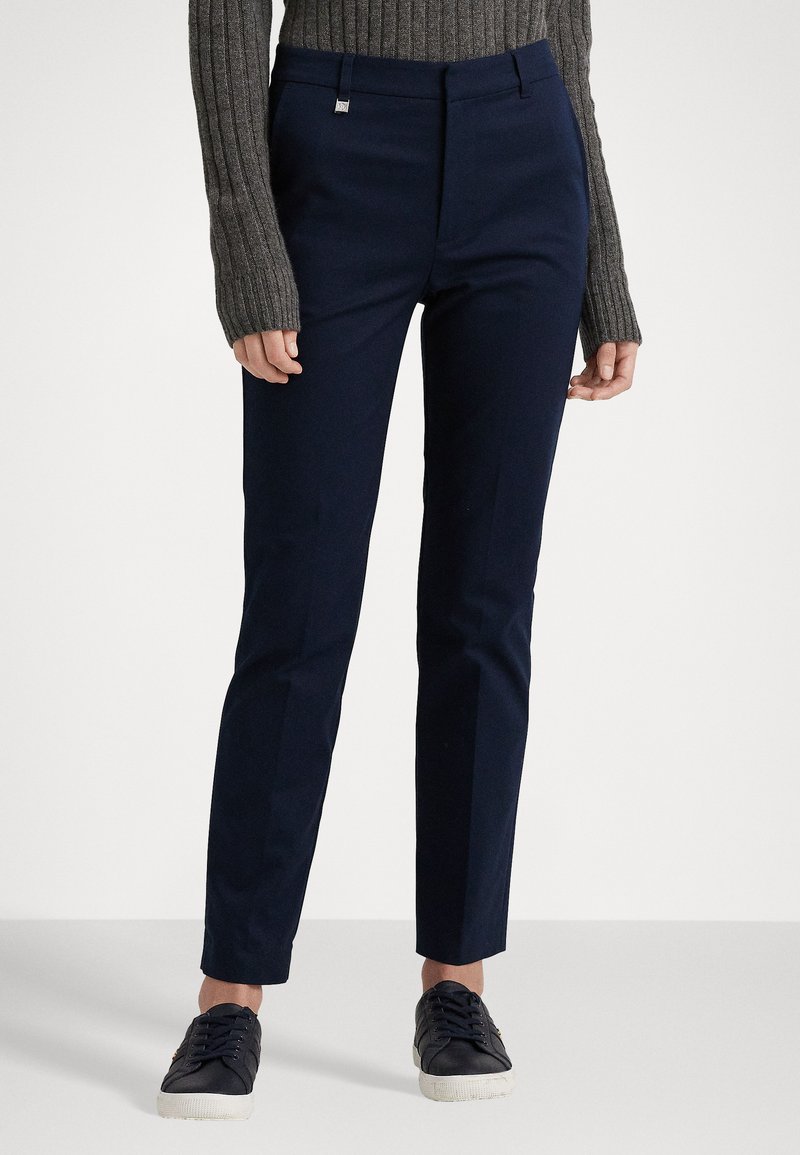 Pantaloni tailored blu navy in tessuto liscio, con frontale piatto e tasche. Abbinati a un maglione grigio a coste e sneakers scure.