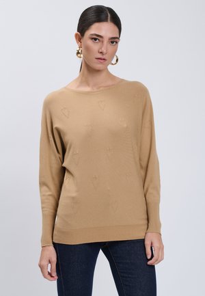 Vrouw met donker haar die een beige trui met subtiele hartjespatronen en donkerblauwe spijkerbroek draagt tegen een effen grijze achtergrond.