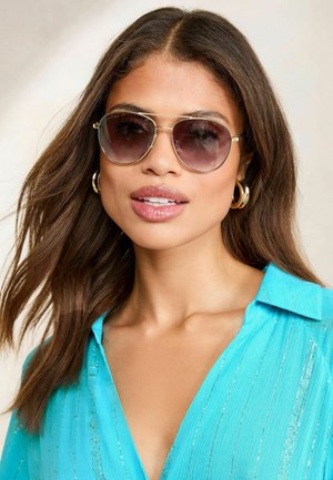 Silberne Sonnenbrille mit grau verlaufenden Gläsern, runder Form, getragen von einem Model in einem türkisfarbenen Oberteil mit vertikalen gestreiften Akzenten.