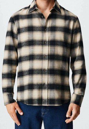 Man draagt een beige en zwart geblokt overhemd met knopen en lange mouwen, gecombineerd met een blauwe spijkerbroek, staand met handen ontspannen langs zijn zij.