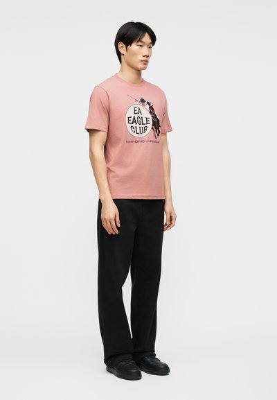 Νεαρός άνδρας που στέκεται, φορώντας ροζ μπλουζάκι με το λογότυπο "EA Eagle Club", μαύρο παντελόνι και μαύρα αθλητικά παπούτσια, σε απλό φωτεινό φόντο.