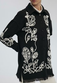 Giacca nera con bottoni, decorata con ricami floreali bianchi, orlo con frange, indossata sopra pantaloni bianchi e una camicia bianca con colletto.