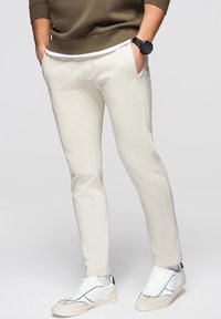 Mand iført beige slim-fit bukser, hvide sneakers og en brun sweatshirt med hænderne i lommerne mod en ensfarvet baggrund.