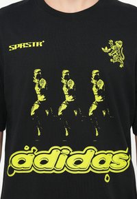 Melnā kokvilnas t-kreklā ir izteiksmīgi dzelteni grafiskie attēli, tostarp trīs figūras un lauvas attēls, ar "adidas" logotipu stilizētā fontā.
