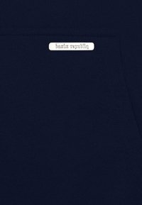 Tissu de sweatshirt bleu marine avec une texture lisse et une étiquette en métal rectangulaire argentée portant l'inscription "bazix republiq" cousue sur la poche.