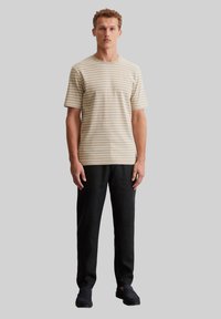 T-shirt beige rayé avec un col rond, associé à un pantalon noir, porté par une personne se tenant contre un fond gris.