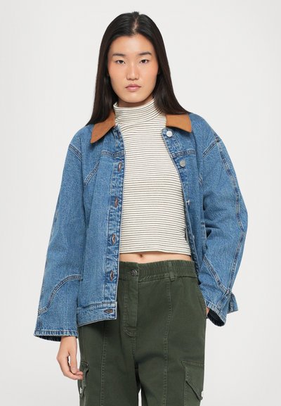 Jeans μπουφάν με καφέ γιακά, μπλε πλύση, κουμπιά μπροστά και μακριά μανίκια. Φοράει πάνω από ένα ριγέ ζιβάγκο και πράσινα cargo παντελόνια.