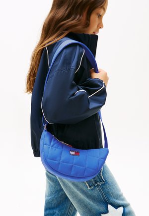 Fată cu jachetă bleumarin și blugi albaștri cu detaliu de stea albă, purtând o geantă albastră matlasată de umăr Tommy Hilfiger.