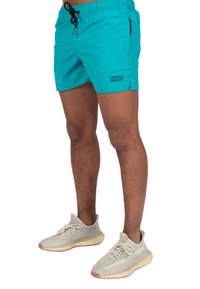 Pantaloni da bagno turchesi con cintura regolabile e tasche laterali, caratterizzati dal logo "WRONG FRIENDS" in nero sul lato sinistro.