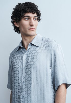 Άνδρας με σγουρά μαύρα μαλλιά φοράει γκρι ανοιχτό πουκάμισο με σχέδιο και κοντά μανίκια, κοιτάζοντας πλάγια σε απλό φόντο.