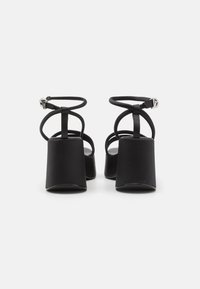Black high-heeled sandals with a matte finish, featuring thin ankle straps and a chunky block heel. Open back design with minimal detailing.