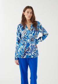 Maglietta a maniche lunghe blu floreale con scollatura a V, con diverse tonalità di blu e bianco, abbinata a pantaloni blu brillanti.