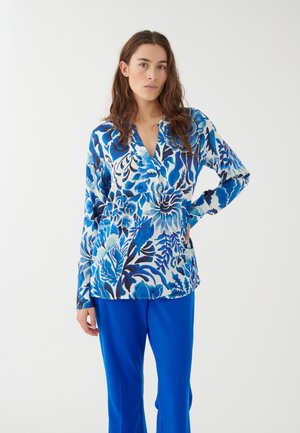 Bluse mit langen Ärmeln in Blau mit V-Ausschnitt, ausgestattet mit verschiedenen Blau- und Weißtönen, kombiniert mit leuchtend blauen Hose.