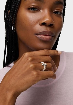 Donna con capelli intrecciati che indossa un anello decorativo di perle e argento sull'indice, con la mano appoggiata vicino al mento, indossando una camicia lavanda.