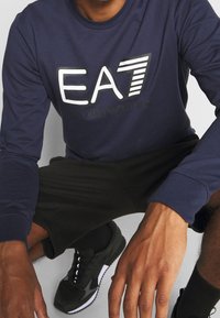 Marinblus med långa ärmar och vit "EA7"-logotyp samt svart text "Emporio Armani"; kombinerat med svarta shorts och svarta träning skor.