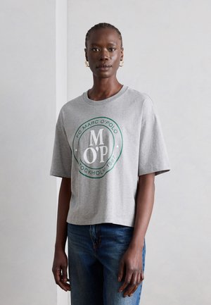 Femeie care poartă un tricou gri Marc O’Polo cu logo circular verde, asortat cu blugi albaștri, stând în fața unui perete simplu, deschis la culoare.