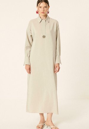 Femme portant une longue robe beige avec col, larges poignets, un bouton décoratif central, des boucles d'oreilles rouges fleuries et des sandales blanches à talons.