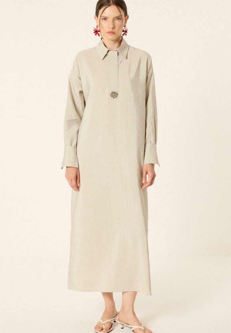 Femme portant une longue robe beige avec col, larges poignets, un bouton décoratif central, des boucles d'oreilles rouges fleuries et des sandales blanches à talons.