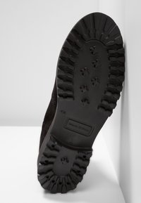 Suola di stivale in suede nero con scanalature profonde, caratterizzata da un logo rettangolare. Il design include una parte superiore in tessuto e una suola in gomma robusta per garantire aderenza.