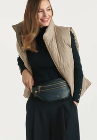 Beżowa kamizelka puchowa, czarny prążkowany golf, granatowe spodnie oraz ciemnozielona nerka z złotymi elementami i podwójnymi kieszeniami na zamek.