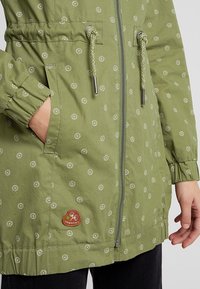 Green jacket made of lightweight fabric, featuring white circular patterns, zipper front, drawstring waist, and elasticated cuffs.