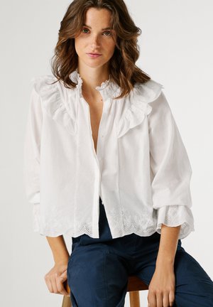 Mujer con cabello castaño ondulado, vestida con una blusa blanca con volantes y bordados en el dobladillo, y pantalones azul oscuro, sentada en un taburete de madera.