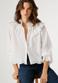 Ženska z valovitimi rjavimi lasmi, oblečena v belo bluzo z volančki in vezenim robom ter temno modre hlače, sedi na lesenem stolčku.