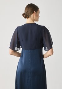 Mørkeblå kjole med transparente fladdermuffesljeder, plissert tekstur på skjørtet og en figurnær overdel, sett fra baksiden.