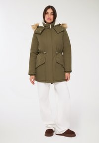 Zielono-oliwkowy płaszcz parka z kapturem z sztucznego futra. Posiada przednie kieszenie, ściągacz w pasie oraz ściągacze na mankietach. Noszony z białymi spodniami i brązowymi kapciami.