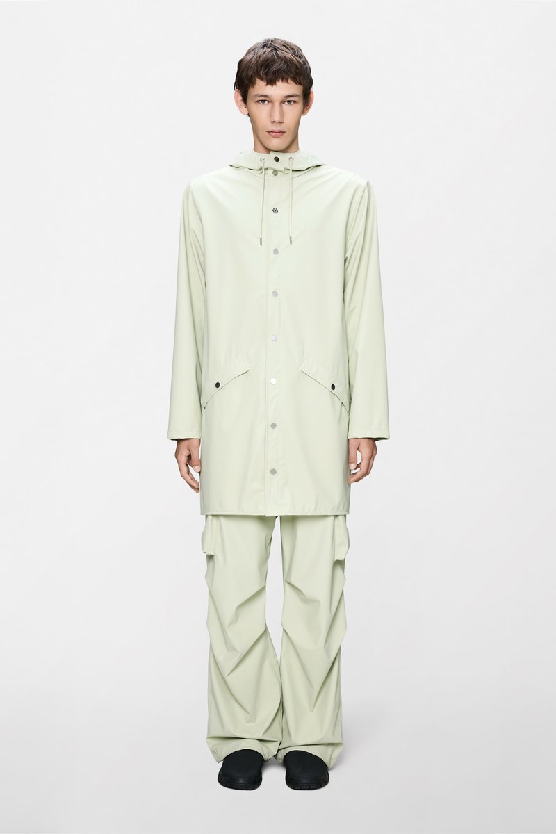 Ανοιχτό πράσινο, αδιάβροχο αδιάβροχο με κουκούλα, κουμπιά κλειδώματος και δύο εμπρόσθιες τσέπες· συνδυασμένο με αντίστοιχο παντελόνι με άνετη εφαρμογή και μαύρα υποδήματα.