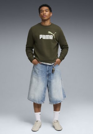 Olivengrøn sweatshirt med hvid "PUMA" logo, båret over en lys skjorte, parret med lyseblå denim shorts og cremede sneakers.