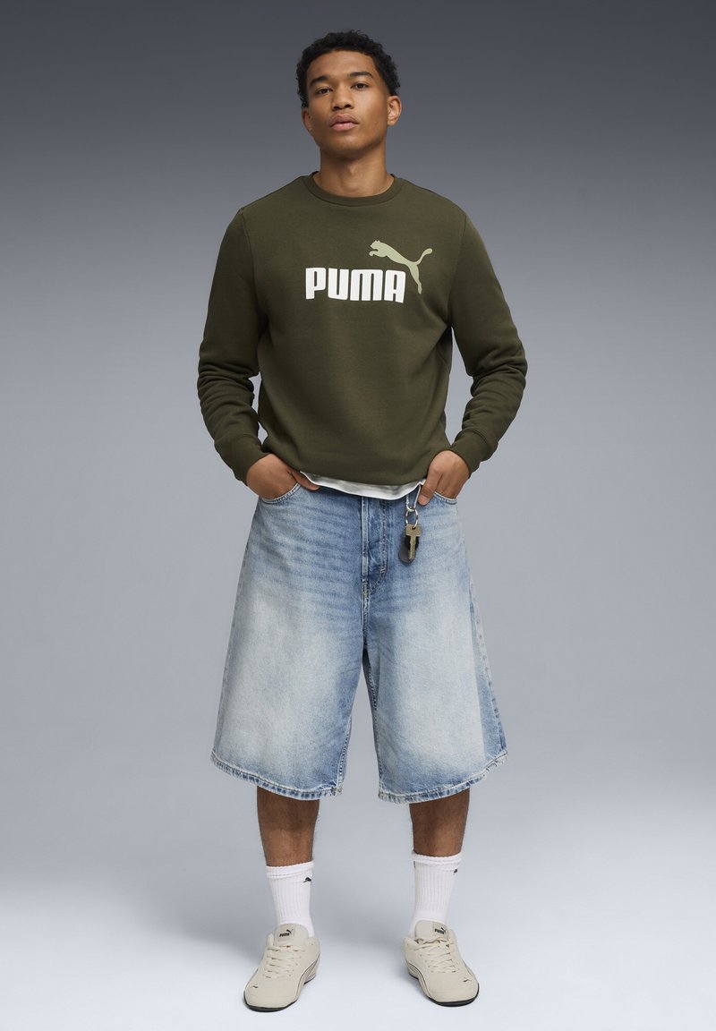 Olivengrøn sweatshirt med hvid "PUMA" logo, båret over en lys skjorte, parret med lyseblå denim shorts og cremede sneakers.