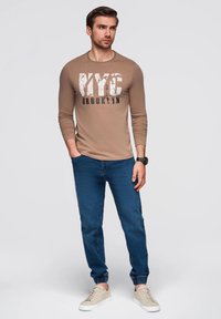 Beige Langarm-T-Shirt mit "NYC BROOKLYN" Grafik, kombiniert mit blauen Joggingjeans und hellbeigen Sneakern, vor einem neutralen Hintergrund stehend.