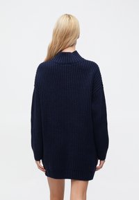 Tumši zils oversize džemperis ar ribotu faktūru, augstu apkakli un garām piedurknēm; tam ir garāks aizmugures apakšmalas apdare un brīvs piegriezums.